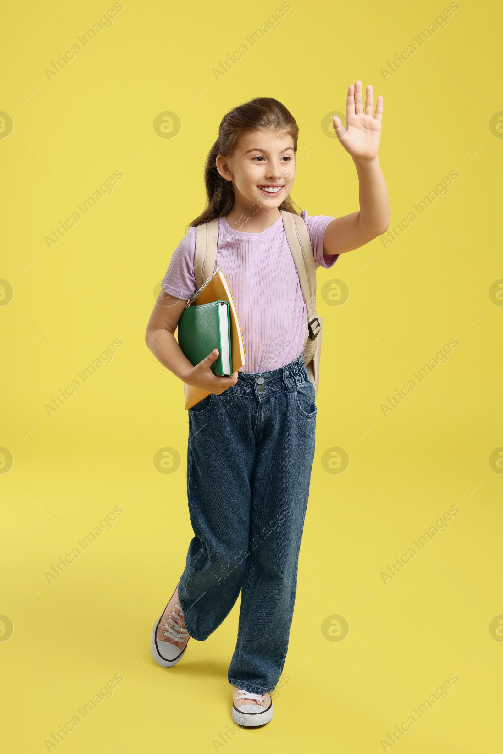 Photo of Girl with backpack and books waving on yellow background