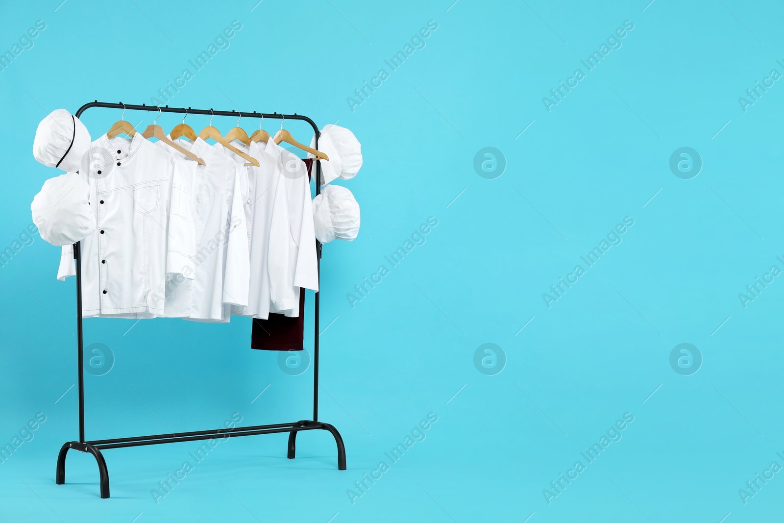 Photo of Different chef's uniforms on clothing rack against light blue background, space for text