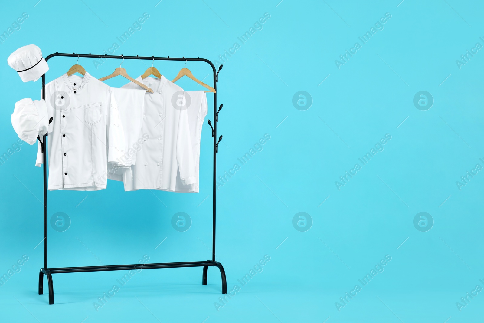 Photo of Different chef's uniforms on clothing rack against light blue background, space for text