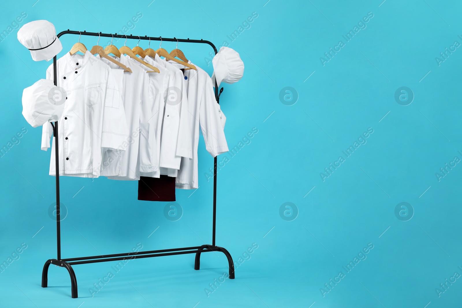 Photo of Different chef's uniforms on clothing rack against light blue background, space for text