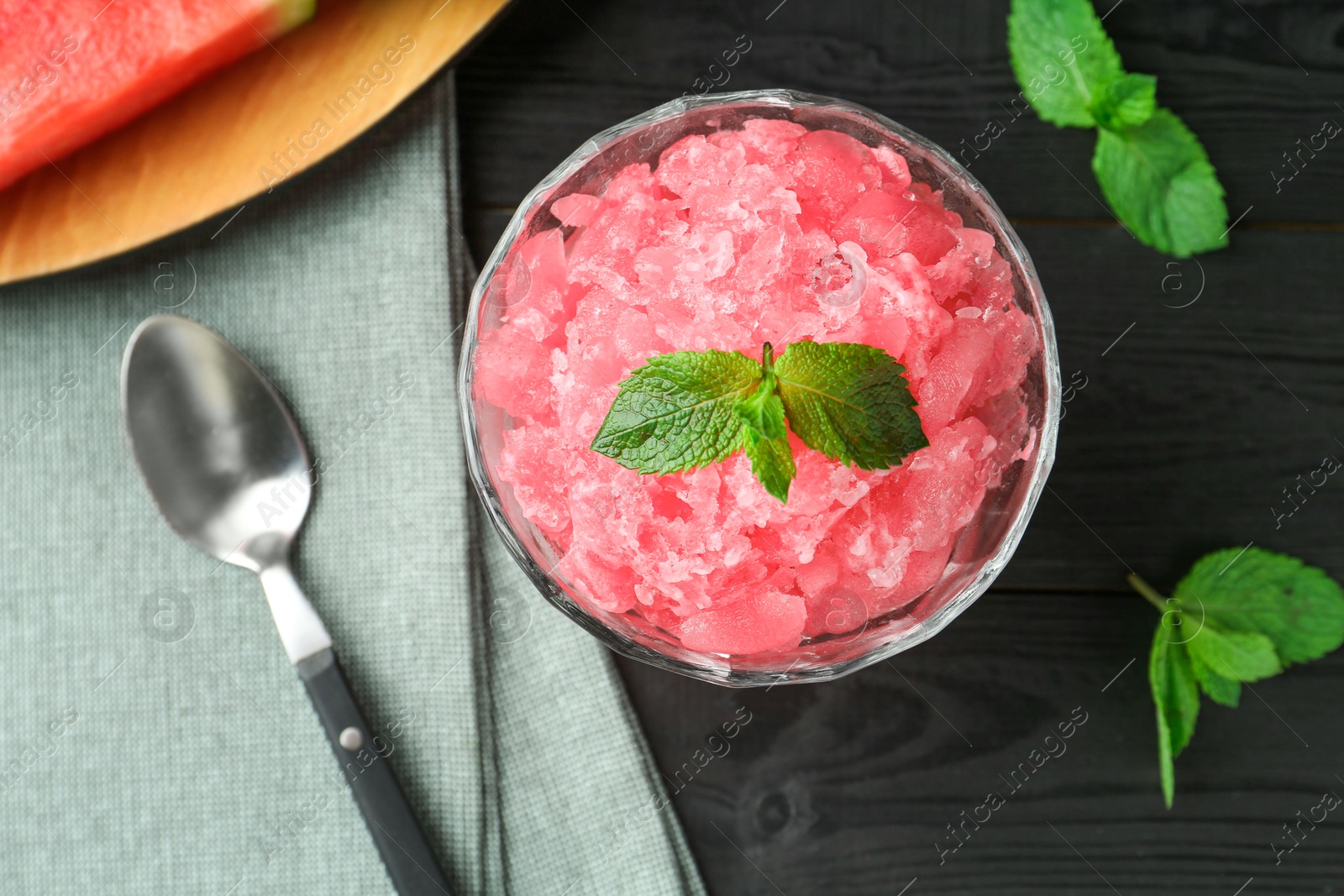 Photo of Tasty watermelon sorbet with mint in glass dessert bowl and spoon on black wooden table, flat lay