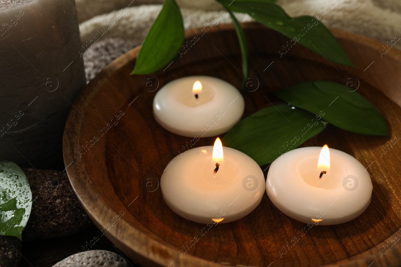 Photo of Burning candles and green leaves in water on table, closeup