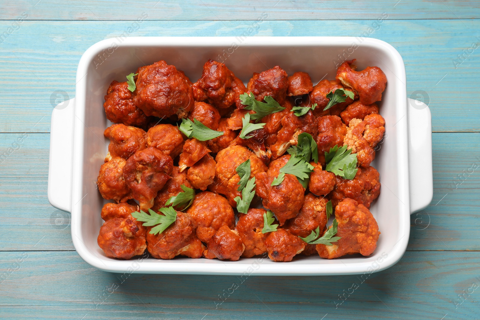 Photo of Baked cauliflower buffalo wings with parsley in baking dish on light blue wooden table, top view