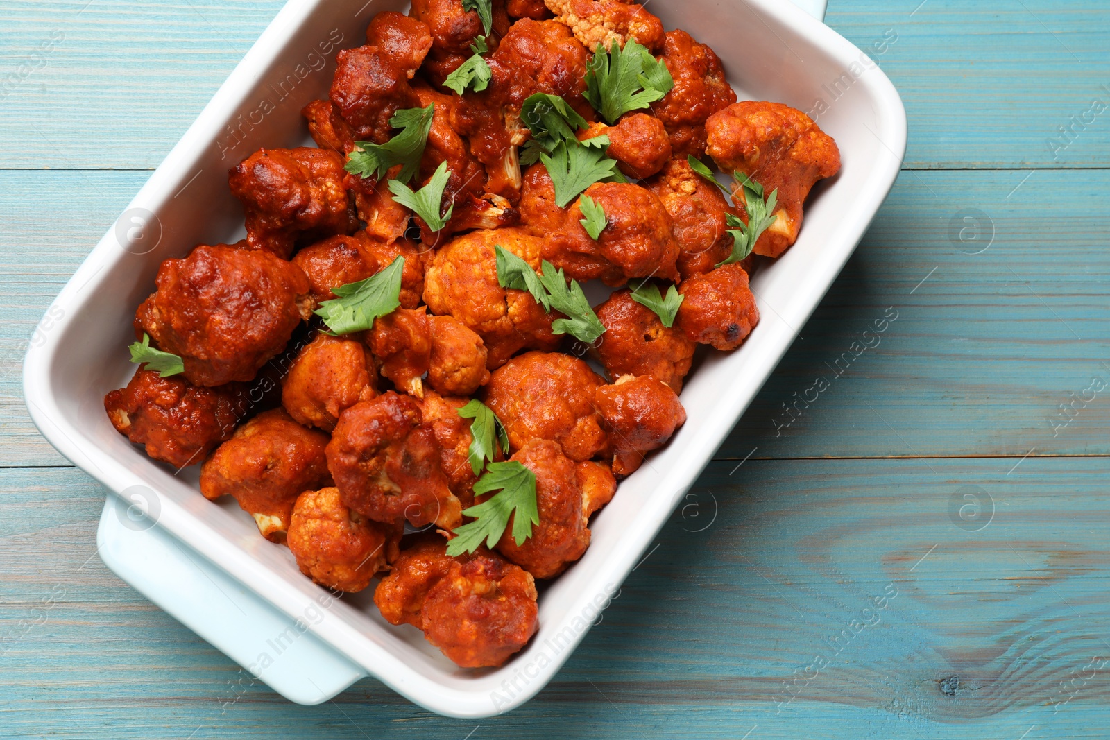 Photo of Baked cauliflower buffalo wings with parsley in baking dish on light blue wooden table, top view