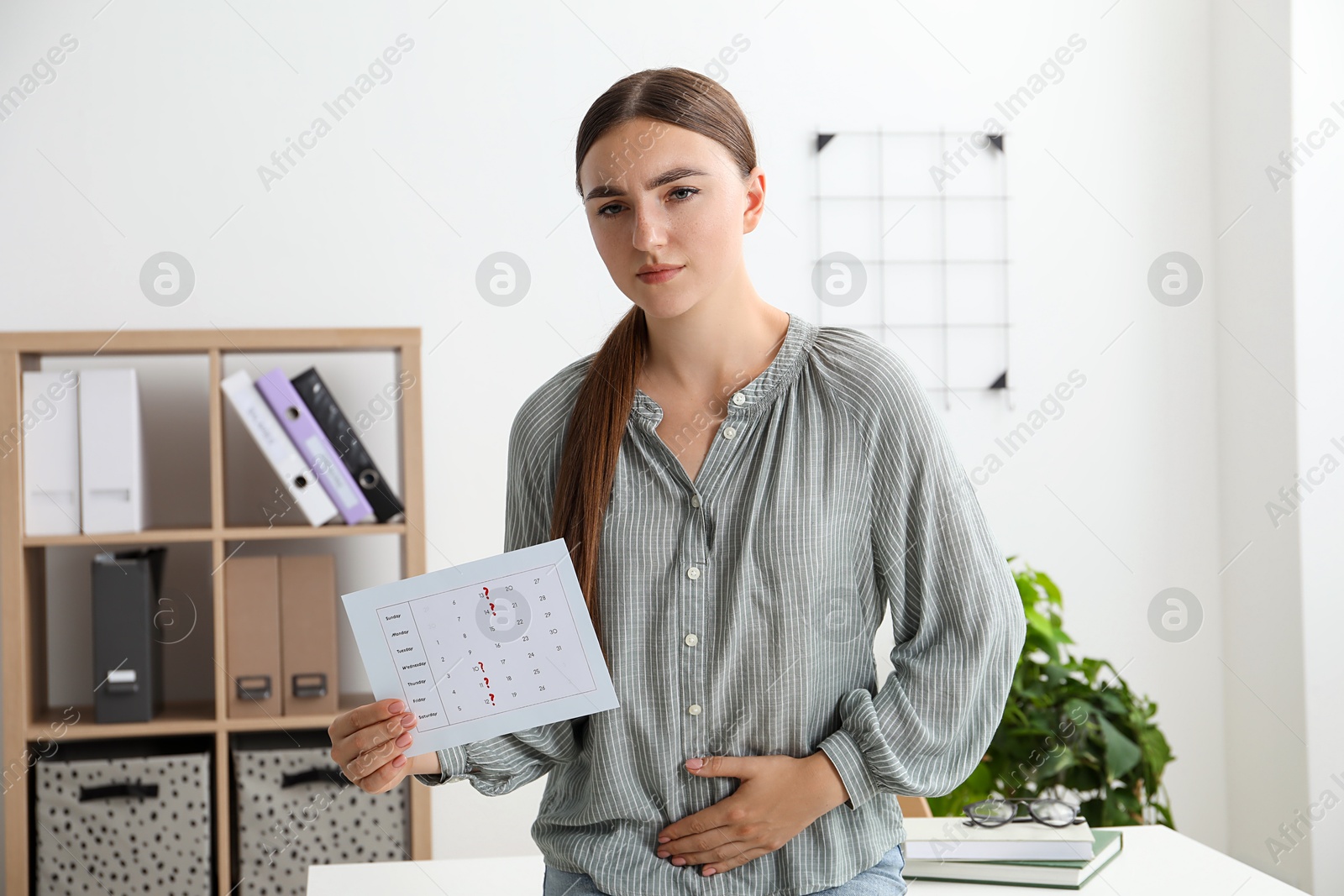 Photo of Woman holding calendar with marked menstrual cycle days and suffering from abdominal pain indoors