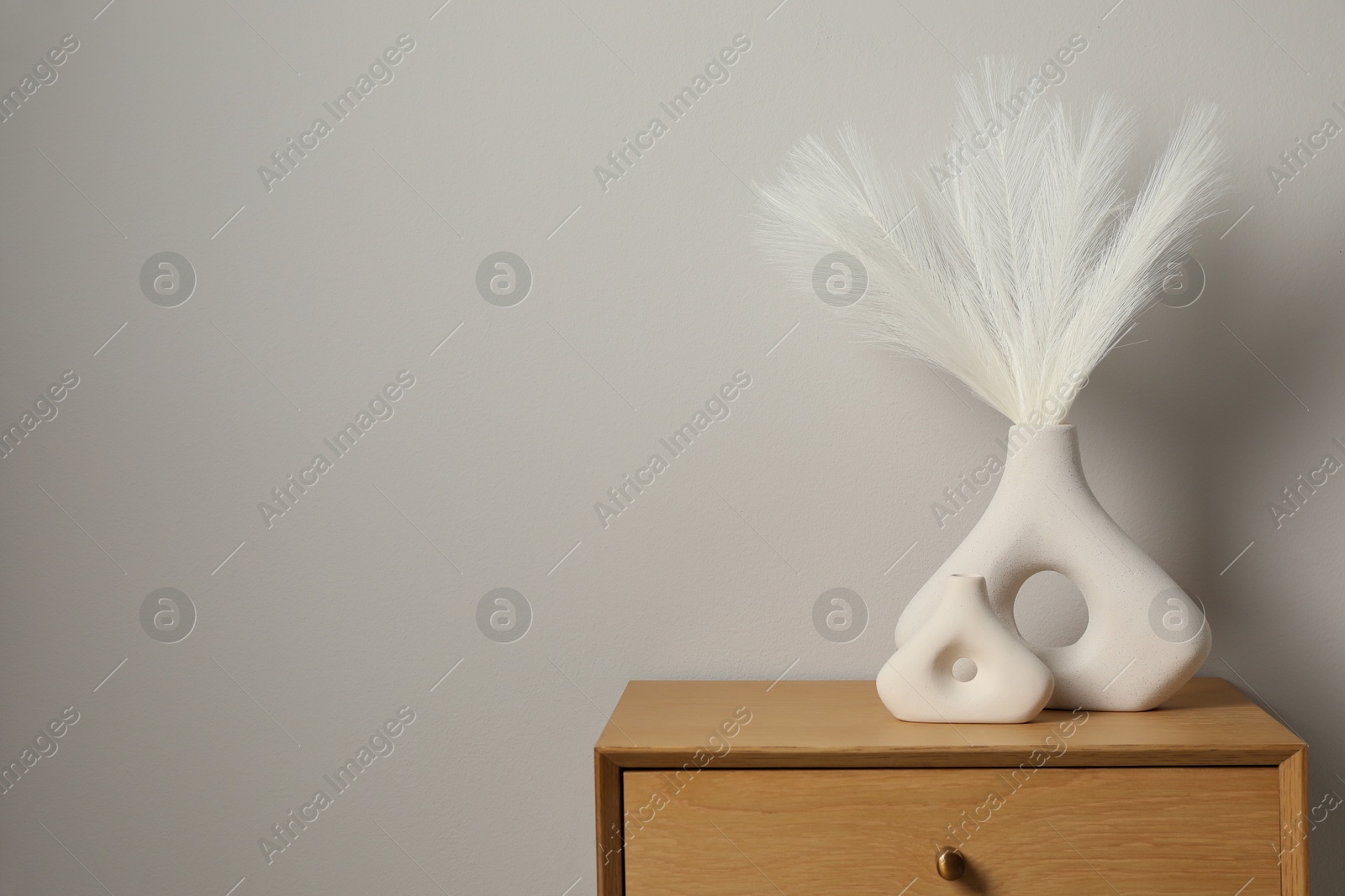 Photo of Ceramic vases with feathers on wooden nightstand near light wall. Space for text