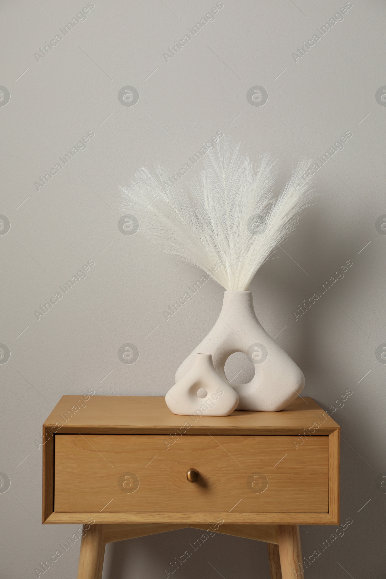 Photo of Ceramic vases with feathers on wooden nightstand near light wall