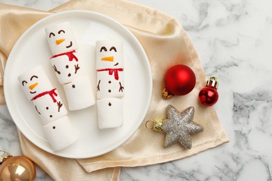 Funny marshmallow snowmen and festive decor on white marble table, top view