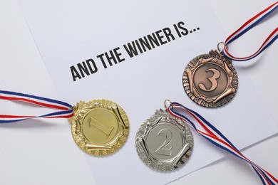 Photo of Sheet of paper with phrase And the winner is and different medals on white table, top view