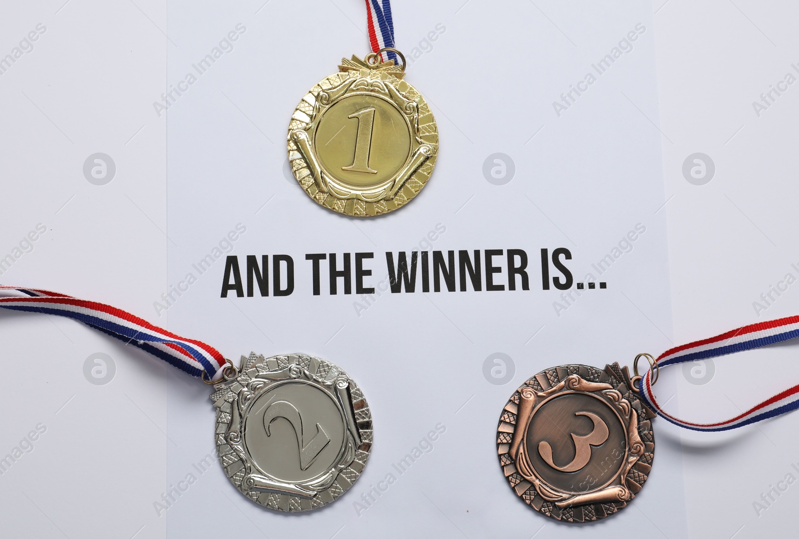 Photo of Sheet of paper with phrase And the winner is and different medals on white table, top view