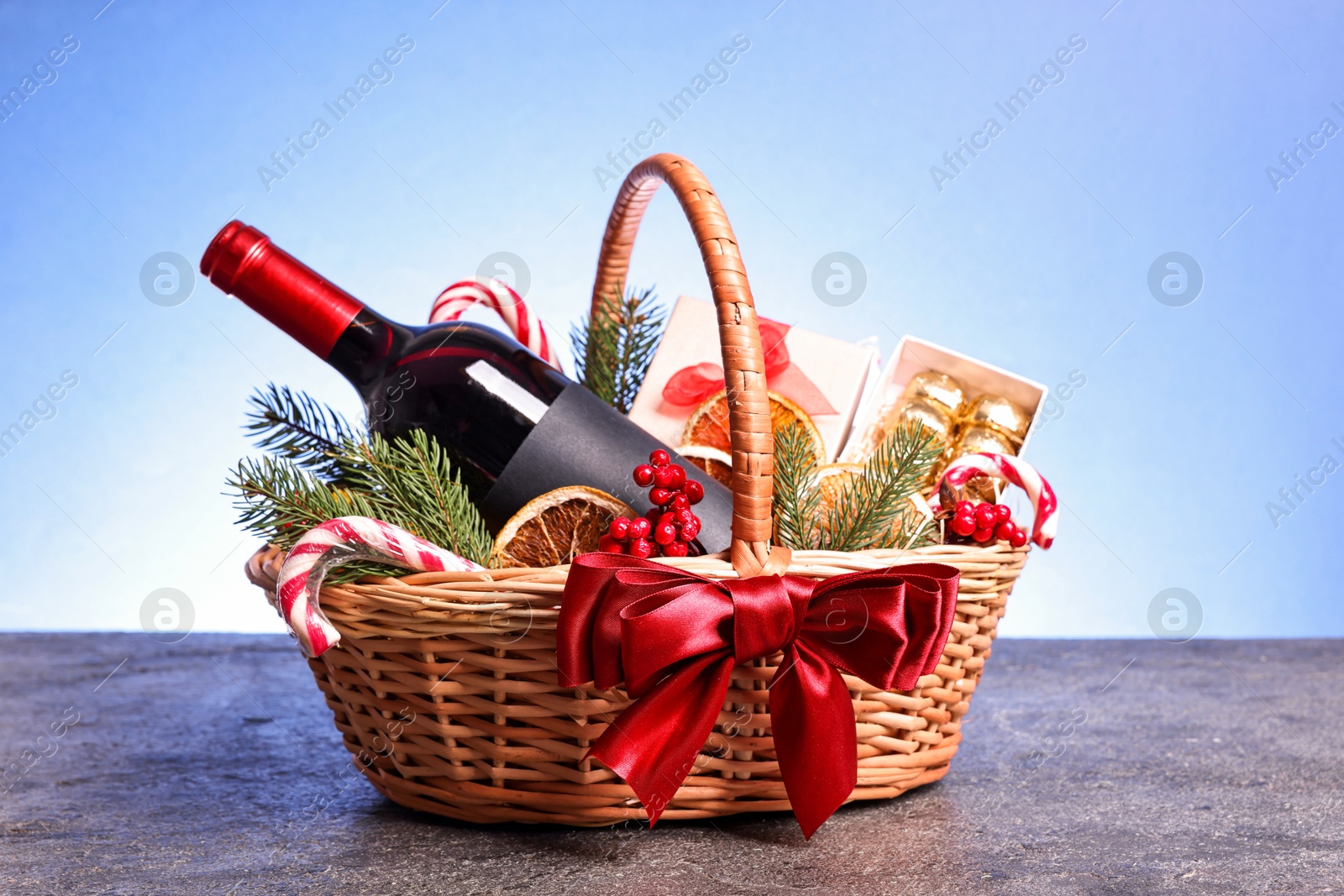 Photo of Wicker gift basket with wine and Christmas decor on grey textured table against light blue background