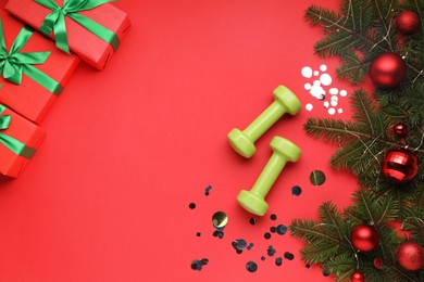 Photo of Dumbbells, fir tree branches, Christmas baubles and gift boxes on red background, flat lay. Space for text