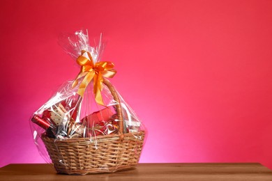 Wicker gift basket with wine on wooden table against pink background, space for text