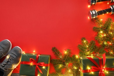 Photo of Dumbbells, fir tree branches, gift boxes and Christmas lights on red background, flat lay. Space for text