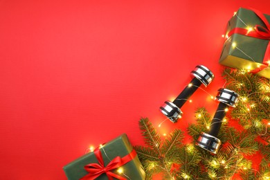 Photo of Dumbbells, fir tree branches, gift boxes and Christmas lights on red background, flat lay. Space for text