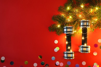 Photo of Dumbbells and fir tree branches with Christmas lights on red background, flat lay. Space for text