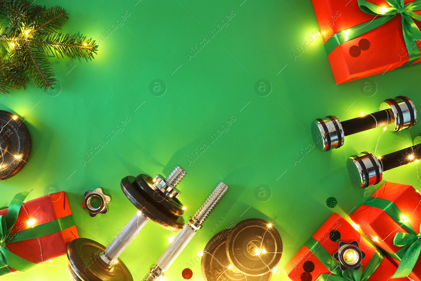 Photo of Dumbbells, fir tree branches, gift boxes and Christmas lights on green background, flat lay. Space for text