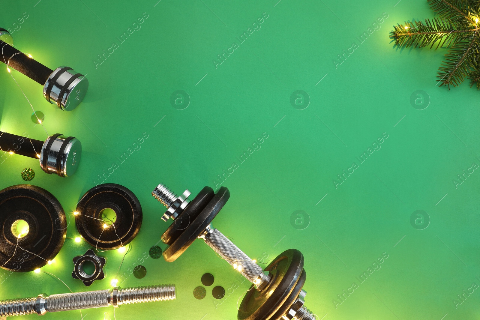 Photo of Dumbbells, fir tree branches and Christmas lights on green background, flat lay. Space for text