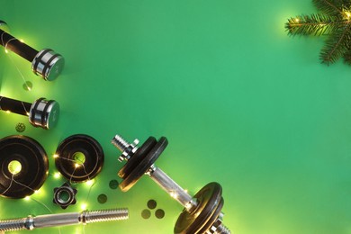 Photo of Dumbbells, fir tree branches and Christmas lights on green background, flat lay. Space for text