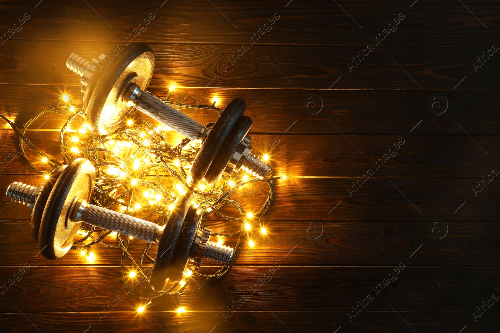 Photo of Dumbbells and Christmas lights on wooden table, top view. Space for text