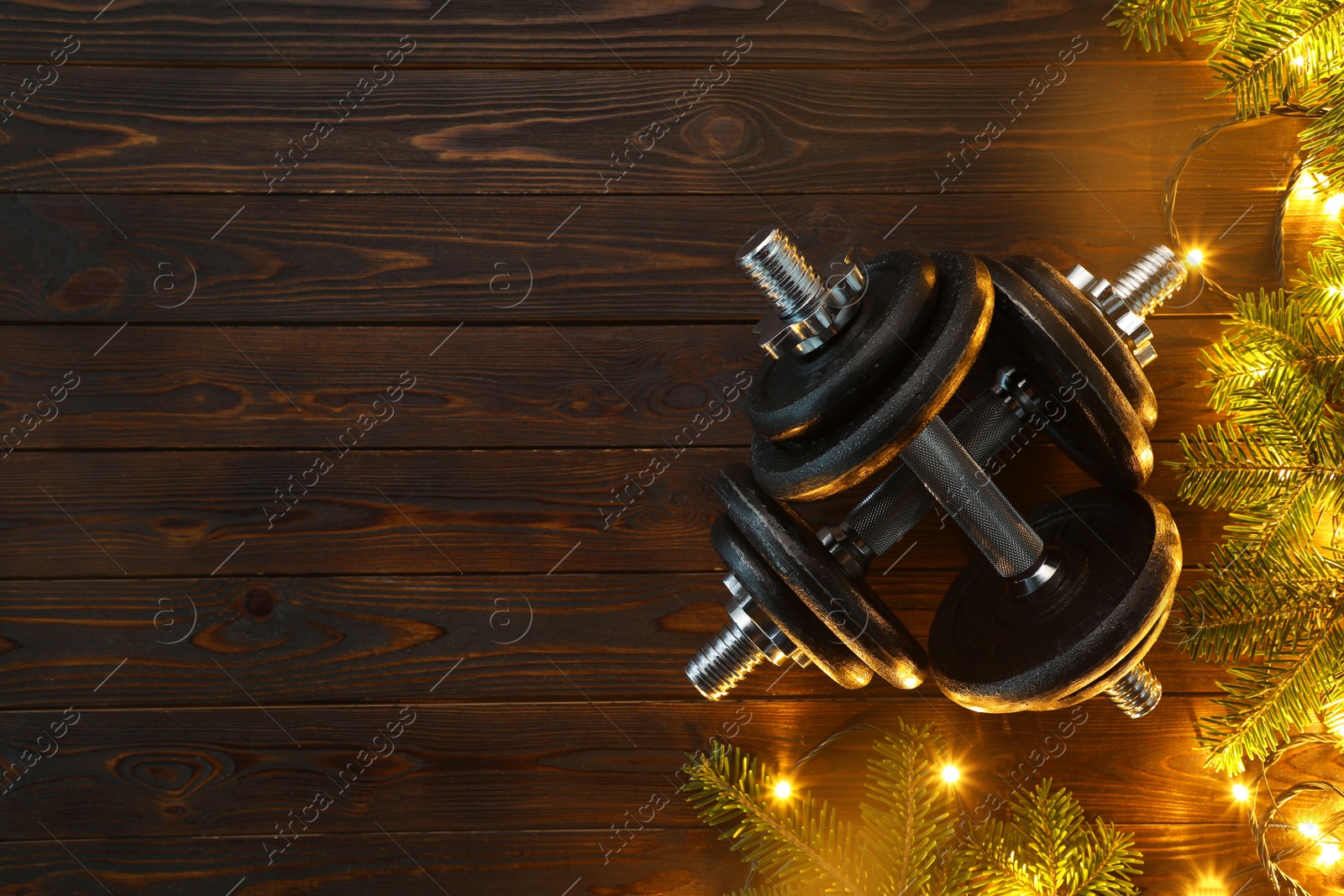 Photo of Dumbbells, fir tree branches and Christmas lights on wooden table, flat lay. Space for text