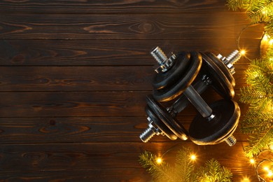 Photo of Dumbbells, fir tree branches and Christmas lights on wooden table, flat lay. Space for text