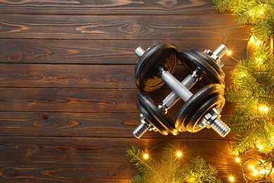 Photo of Dumbbells, fir tree branches and Christmas lights on wooden table, flat lay. Space for text