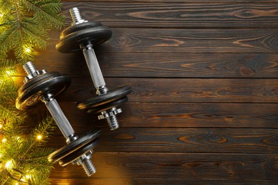 Photo of Dumbbells, fir tree branches and Christmas lights on wooden table, flat lay. Space for text