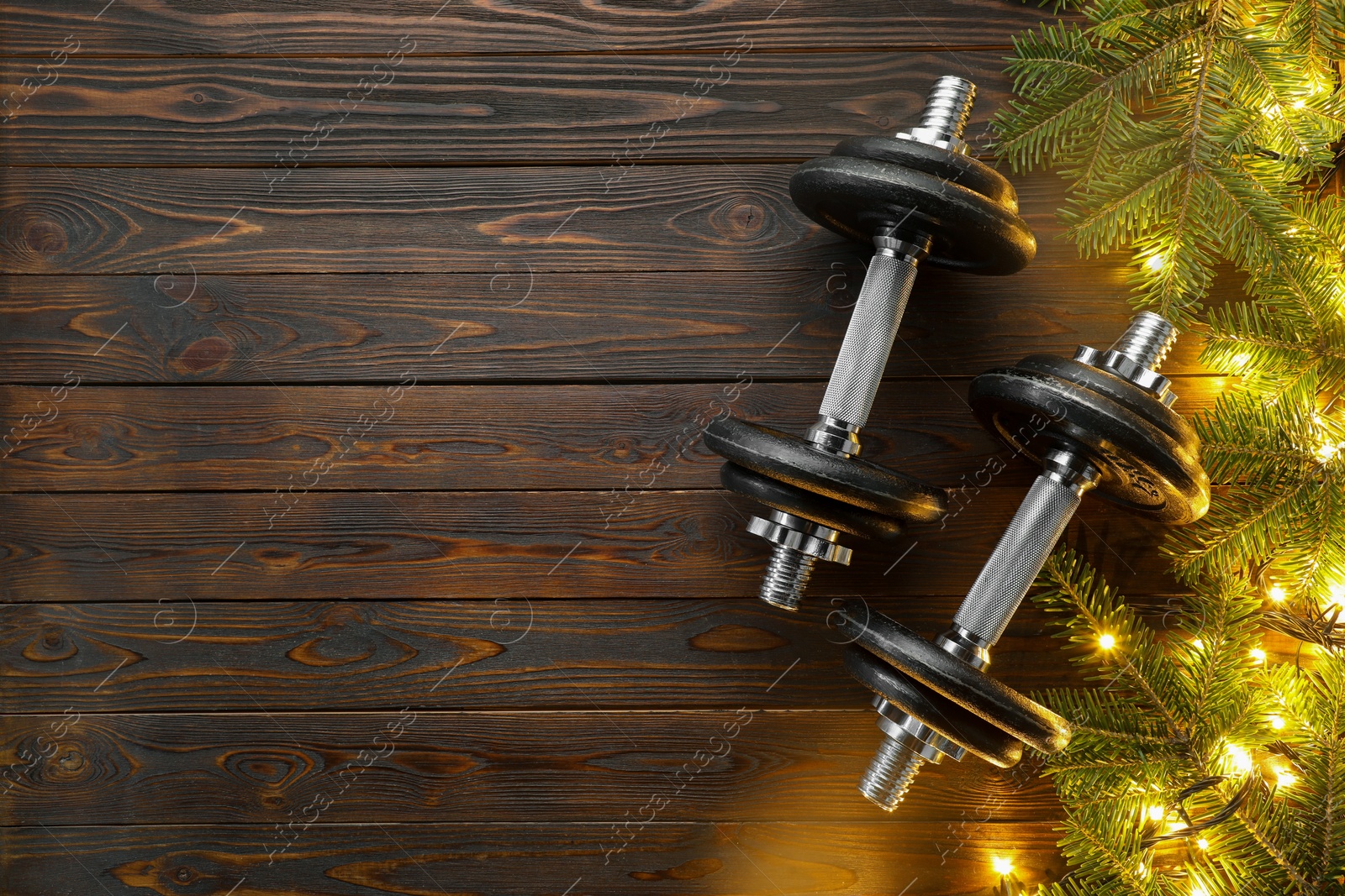 Photo of Dumbbells, fir tree branches and Christmas lights on wooden table, flat lay. Space for text