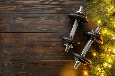 Photo of Dumbbells, fir tree branches and Christmas lights on wooden table, flat lay. Space for text