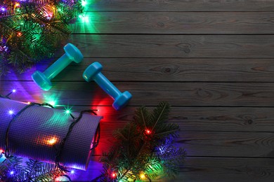 Photo of Dumbbells, mat, fir tree branches and Christmas lights on wooden table, flat lay. Space for text