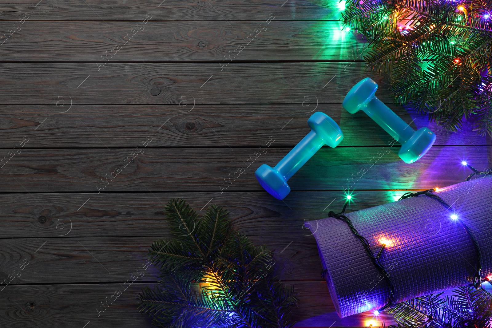 Photo of Dumbbells, mat, fir tree branches and Christmas lights on wooden table, flat lay. Space for text