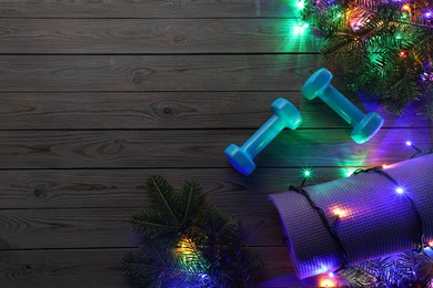 Photo of Dumbbells, mat, fir tree branches and Christmas lights on wooden table, flat lay. Space for text