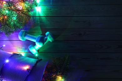 Photo of Dumbbells, mat, fir tree branches and Christmas lights on wooden table, flat lay. Space for text