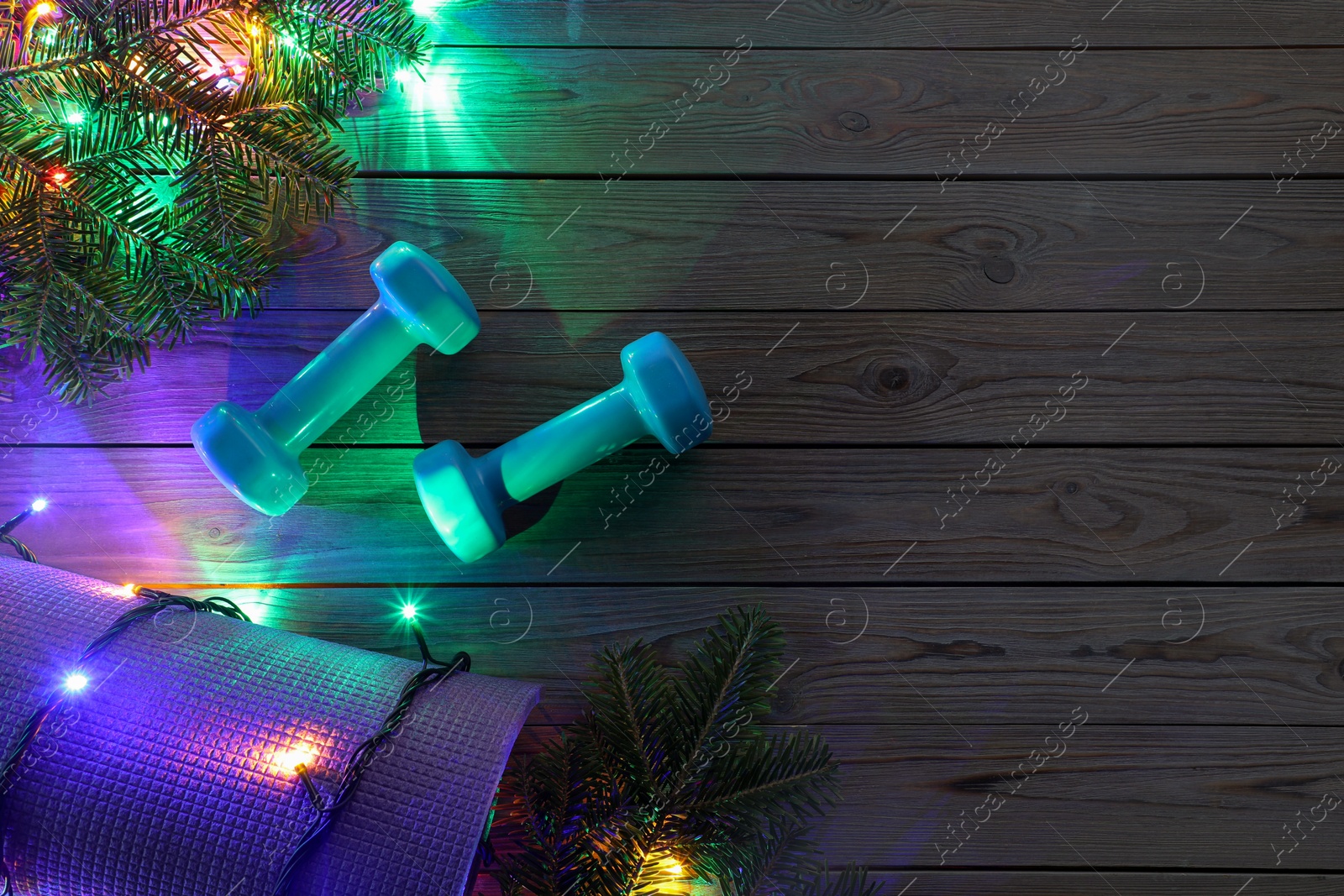 Photo of Dumbbells, mat, fir tree branches and Christmas lights on wooden table, flat lay. Space for text