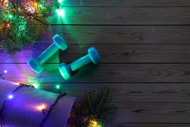 Photo of Dumbbells, mat, fir tree branches and Christmas lights on wooden table, flat lay. Space for text