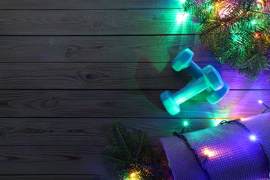 Photo of Dumbbells, mat, fir tree branches and Christmas lights on wooden table, flat lay. Space for text