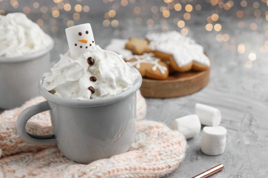 Photo of Funny snowman made with marshmallow and whipped cream in cup of hot drink served on grey background against Christmas lights, closeup