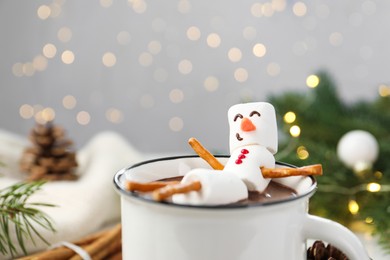 Photo of Funny snowman made with marshmallows in cup of hot chocolate and winter decor against Christmas lights, closeup