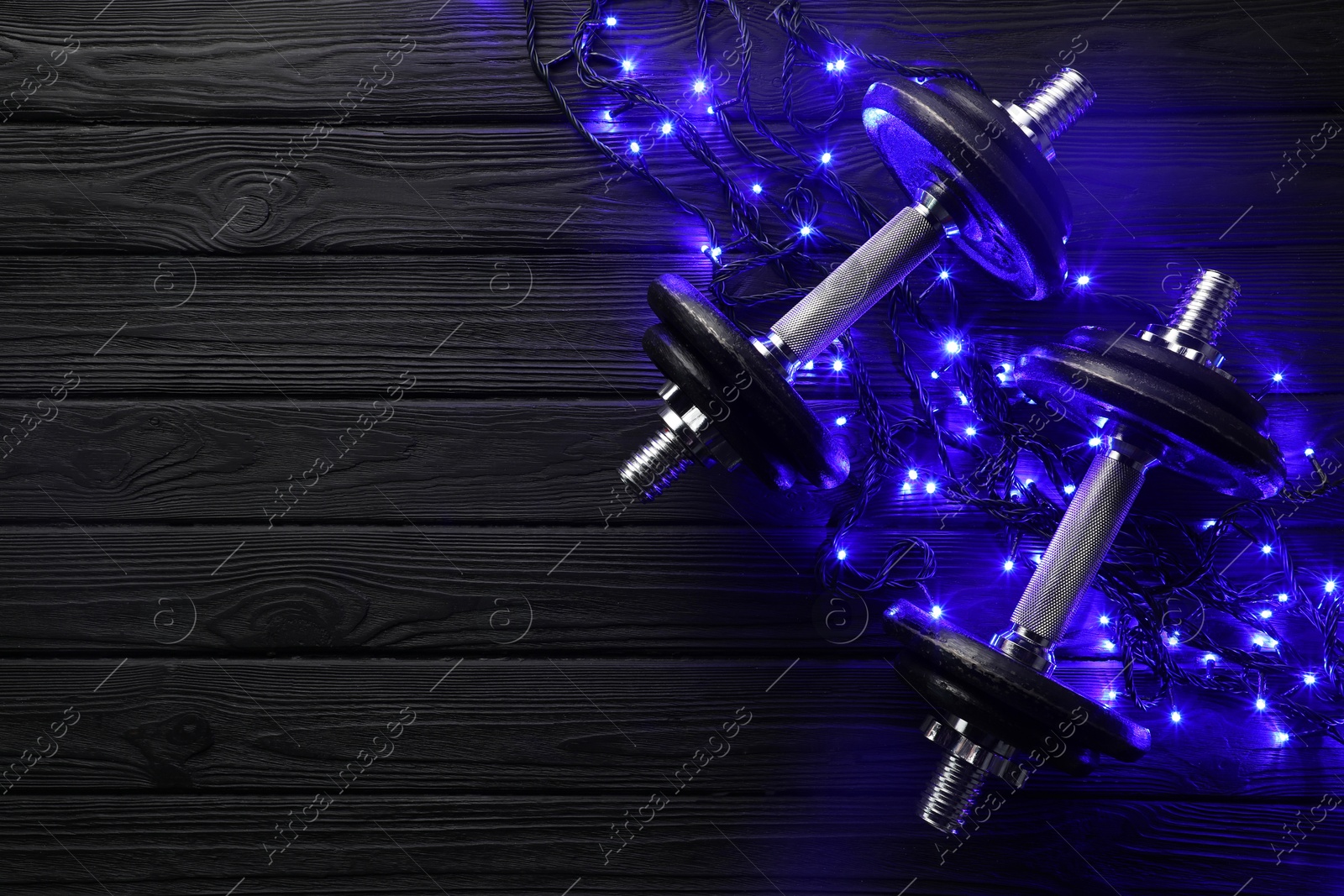 Photo of Dumbbells and Christmas lights on wooden table, flat lay. Space for text