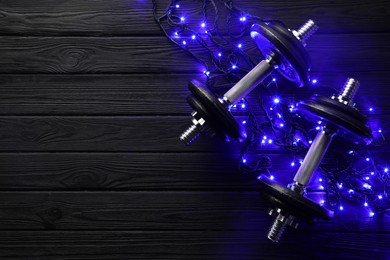 Photo of Dumbbells and Christmas lights on wooden table, flat lay. Space for text