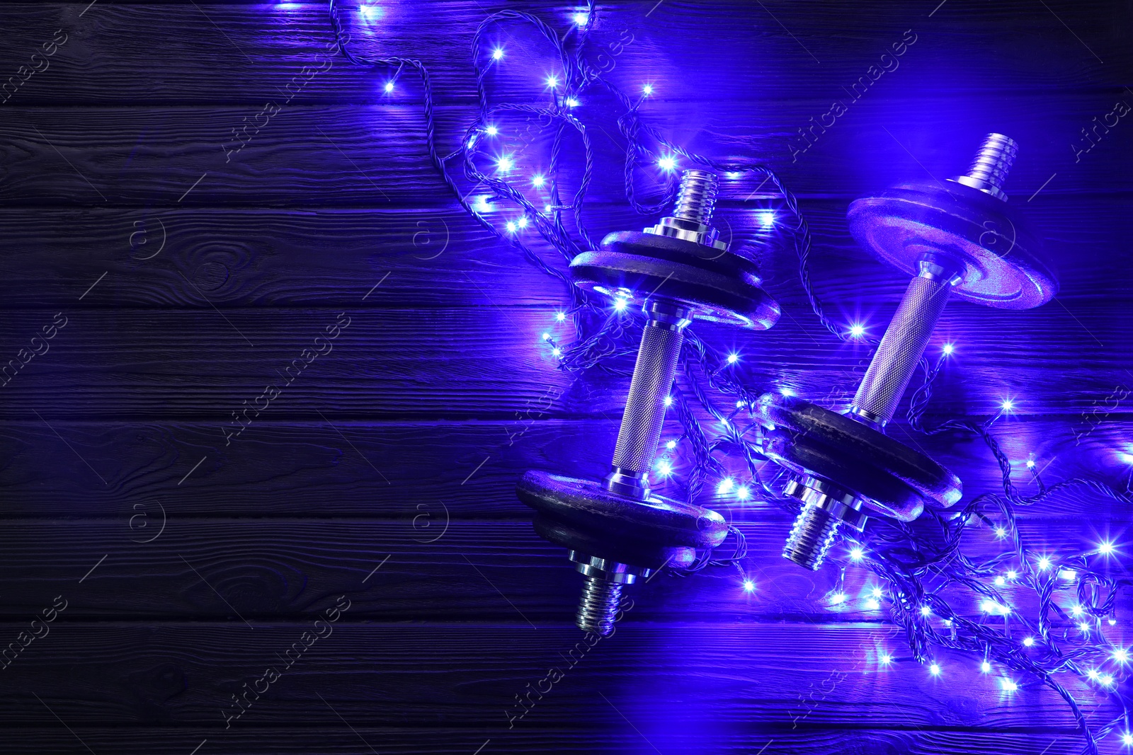 Photo of Dumbbells and Christmas lights on wooden table, flat lay. Space for text