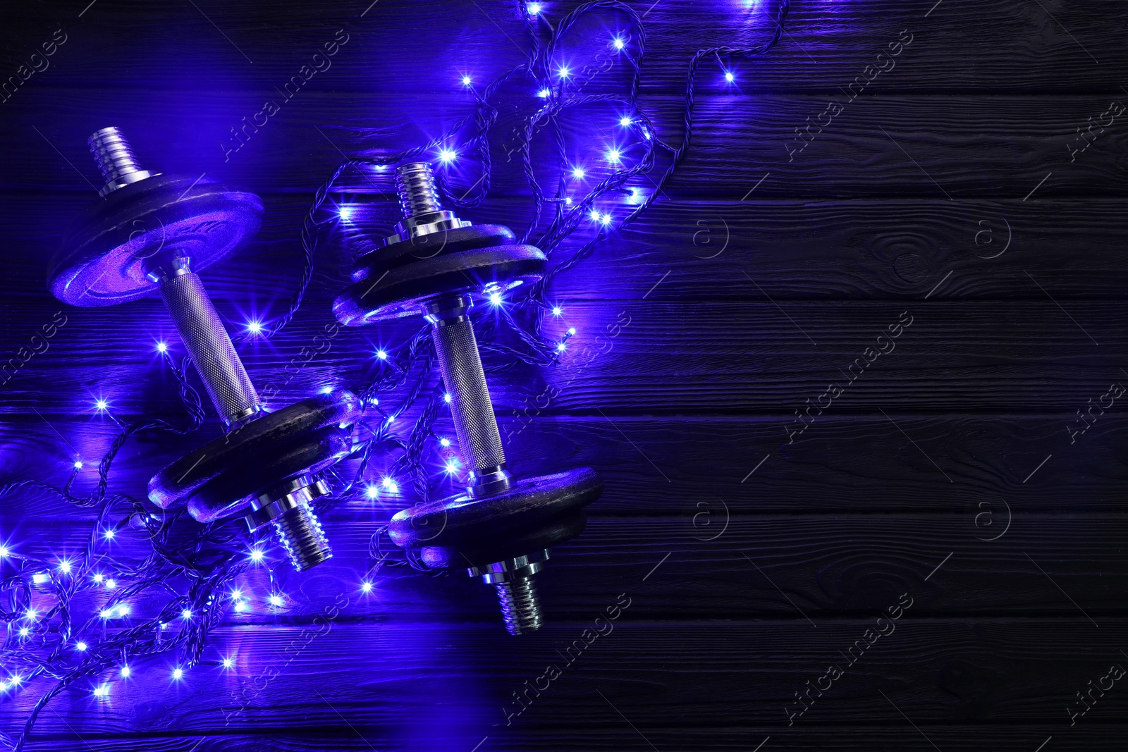 Photo of Dumbbells and Christmas lights on wooden table, flat lay. Space for text