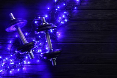 Photo of Dumbbells and Christmas lights on wooden table, flat lay. Space for text