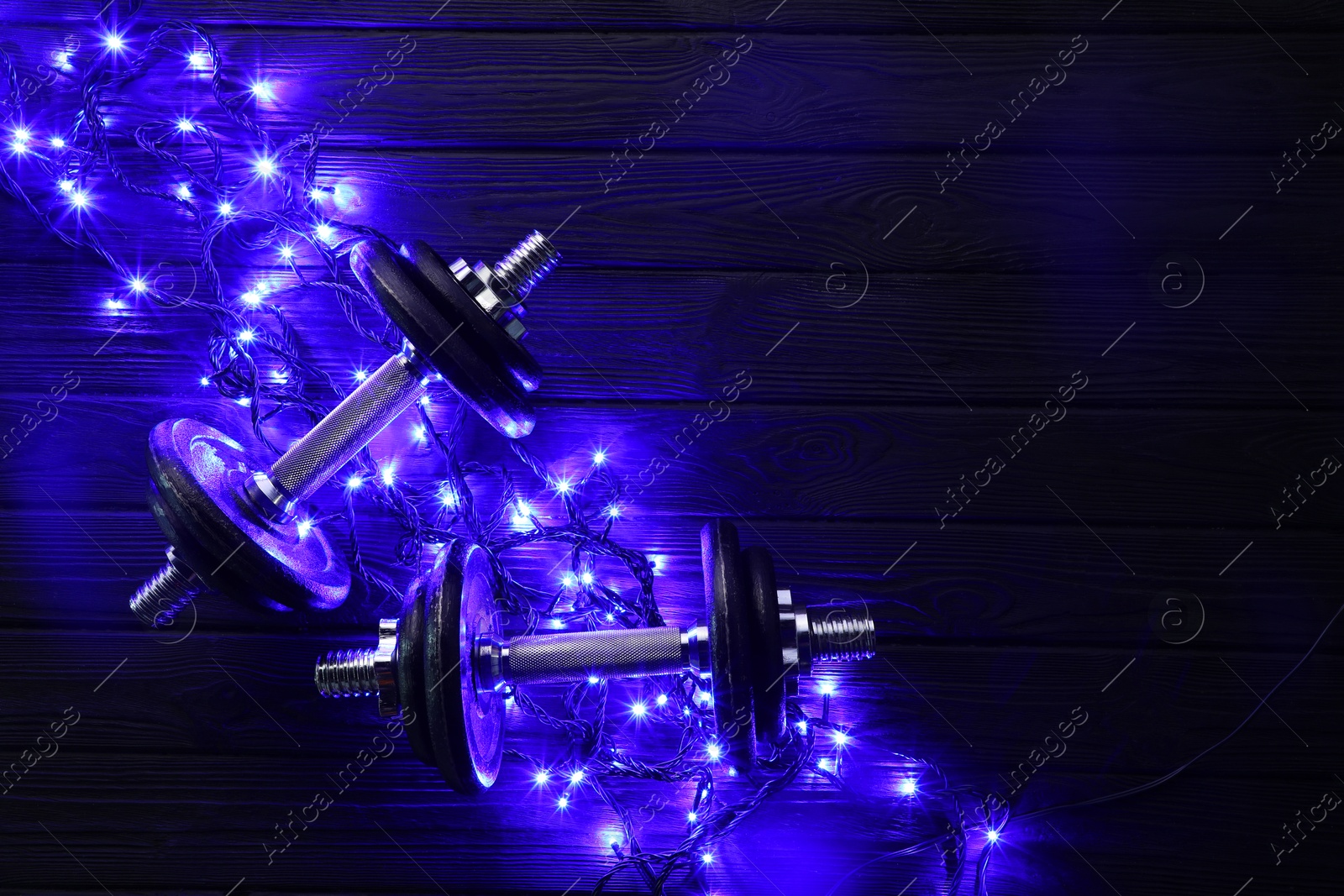 Photo of Dumbbells and Christmas lights on wooden table, flat lay. Space for text