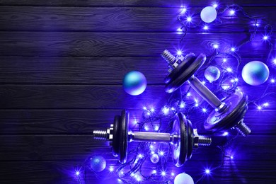 Dumbbells, baubles and Christmas lights on wooden table, flat lay. Space for text