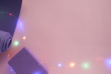 Photo of Yoga mat, block and Christmas lights on pink background, flat lay. Space for text