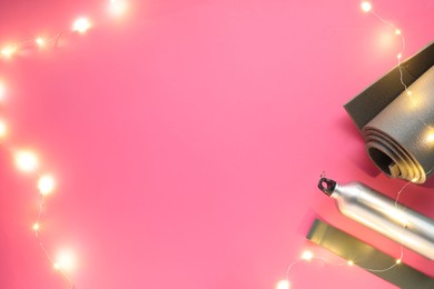 Photo of Yoga mat, band, bottle and Christmas lights on pink background, flat lay. Space for text