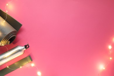 Photo of Yoga mat, band, bottle and Christmas lights on pink background, flat lay. Space for text