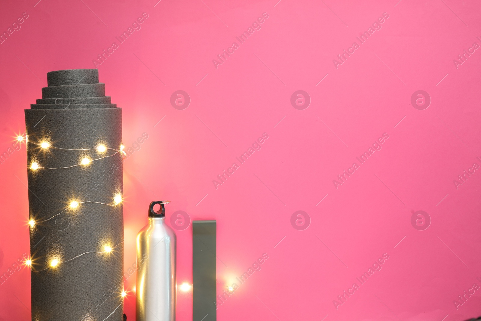 Photo of Yoga mat, band, bottle and Christmas lights on pink background, flat lay. Space for text